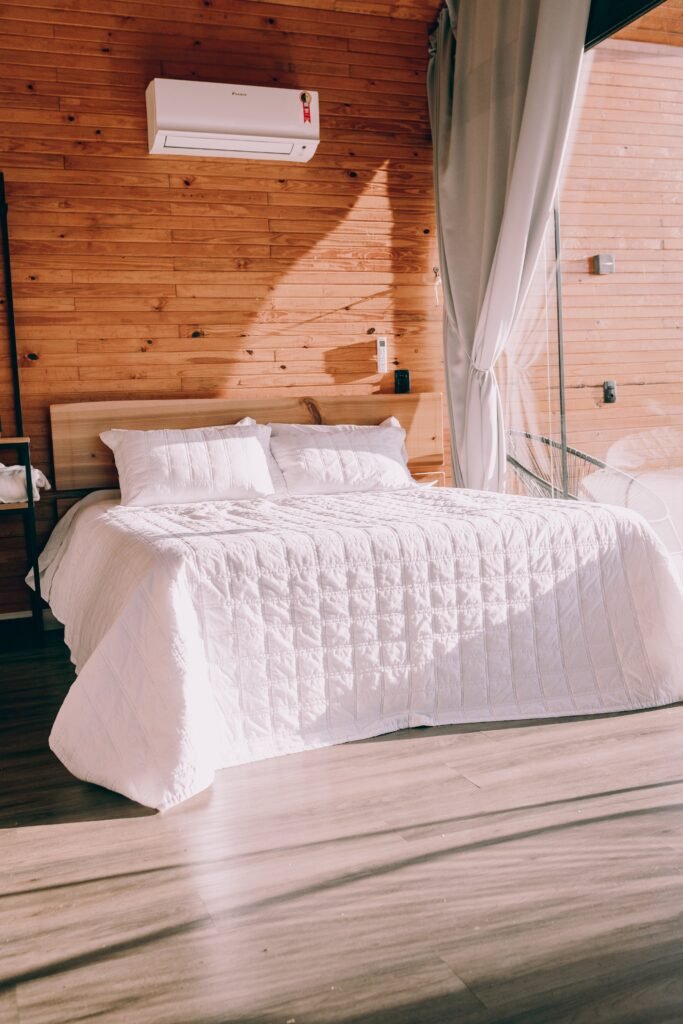 Cama doble con un cubrecolchon blanco y ropa de cama limpia en un dormitorio moderno con paredes de madera y luz natural.