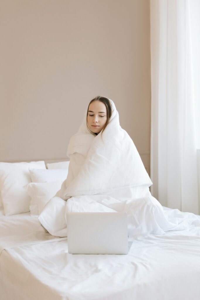 A woman wrapped in a white blanket sits on a bed using a laptop, indoors.