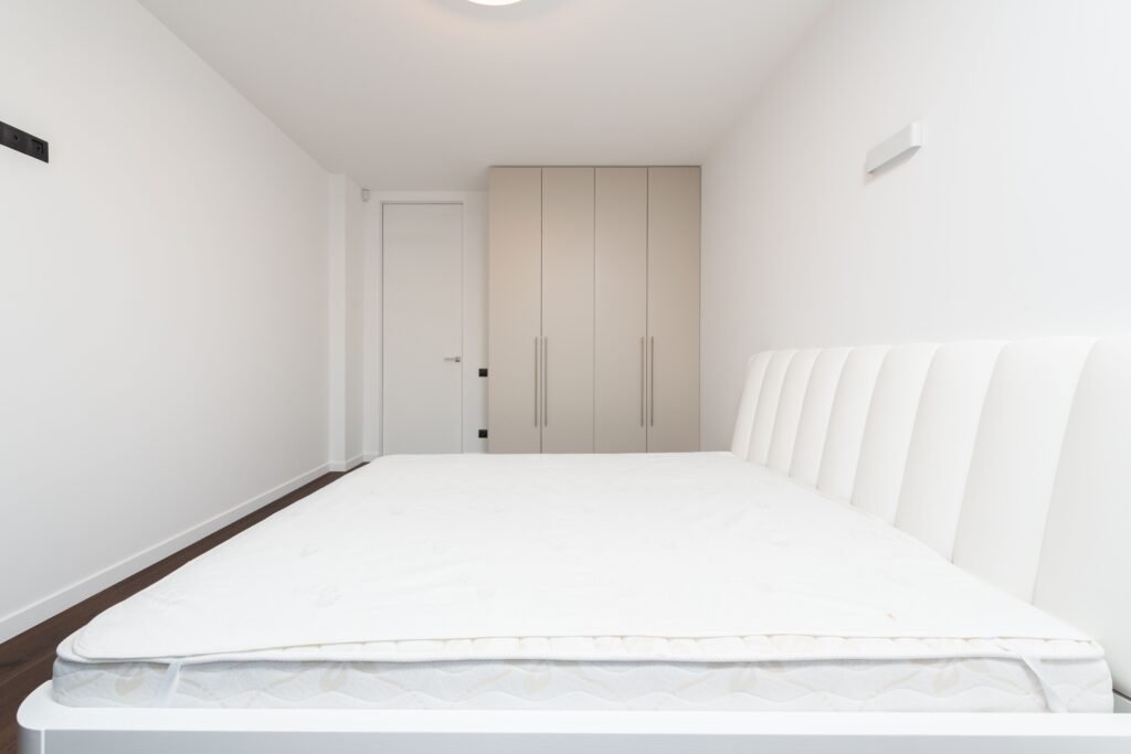 Alt Text: "A luxurious contemporary minimalist bedroom featuring a clean-lined wooden bed with a soft white mattress topper, a sleek integrated wardrobe, and warm recessed LED lighting. This serene design showcases the type of comfort and style highlighted in our list of the 5 Best Mattress Toppers for 2026.