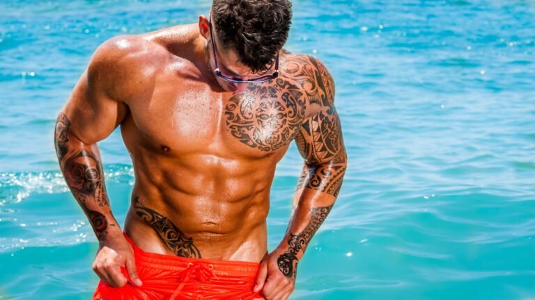 Hombre musculoso con tatuaje en el pecho posando en el agua, ideal para mostrar los resultados de una crema depiladora para hombres.